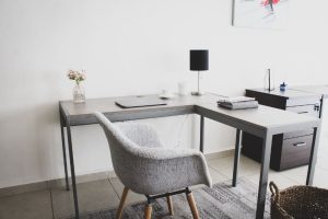 Apartments in Farragut A modern home office with a gray desk, a gray upholstered chair, black lamp, laptop, notebooks, and a vase of pink flowers, set against a white wall with minimalist art featuring an