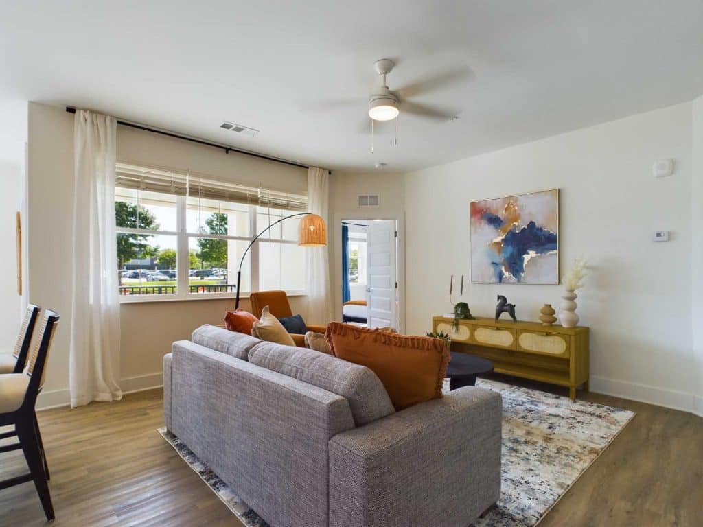 Apartments in Farragut A modern living room with a gray sofa, orange pillows, a wooden sideboard, and a floor lamp. Large window with white curtains and a ceiling fan overhead. Artwork and decorative items on the sideboard.