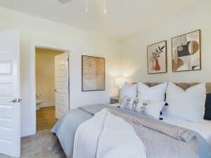 Apartments in for rent Farragut TN A modern bedroom with a neatly made bed, pillows, and artwork on the wall. An open door reveals a bathroom with a toilet. The room is well-lit and decorated in neutral tones. Anchor 934 Apartments in Biddle Farms Town Center