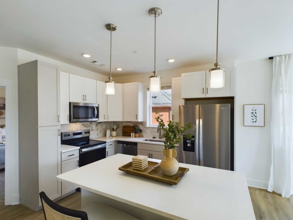 Apartments in Farragut Modern kitchen with white cabinets, stainless steel appliances, and pendant lights hanging over a white island with a small tray and vase. Natural light streams in from a window over the sink.