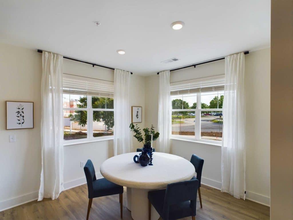 Apartments in Farragut A corner room with large windows, white curtains, a round white table with a blue vase and green branches, and four blue chairs.