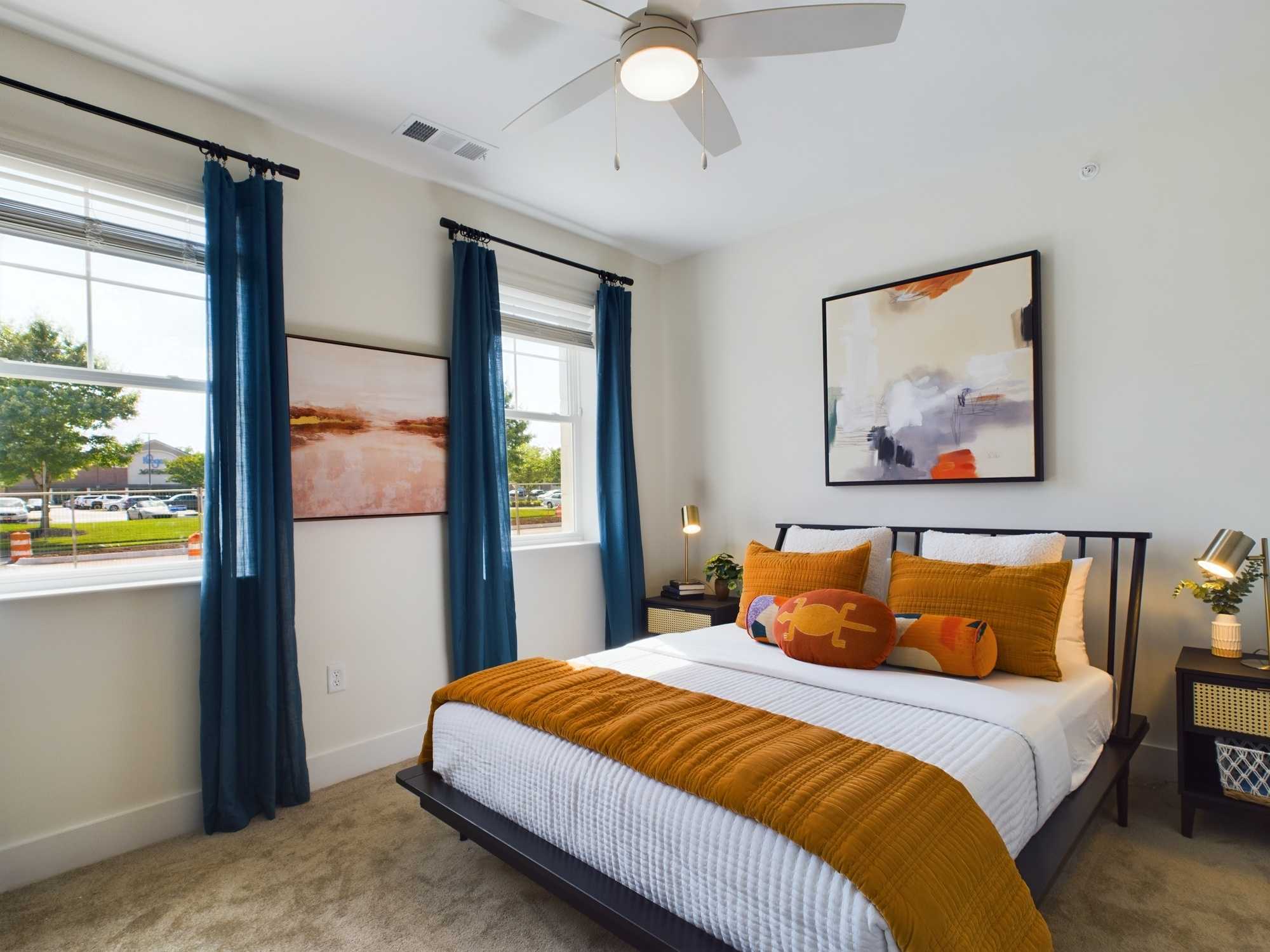 Apartments in Farragut A bedroom with a bed, white sheets, and orange accents. Two abstract paintings hang on the walls, and two windows with blue curtains allow natural light to enter the room. A ceiling fan is on the ceiling.