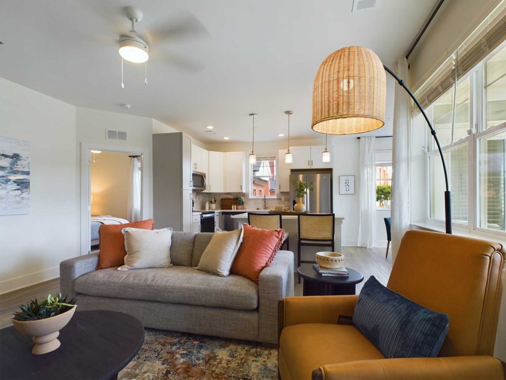 Apartments in Farragut Modern living room with a beige sofa, colorful cushions, brown armchair, and a floor lamp. The open-plan space includes a kitchen with pendant lights and a dining area. A bedroom is visible through an open door.