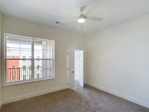 Apartments in Farragut A bright, empty room with beige carpet, a ceiling fan, a large window, and a white door that is slightly open—an inviting space in one of our premium rental properties.