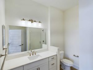 Apartments in Farragut A modern bathroom in a rental apartment features a large mirror, a sink with cabinets below, a toilet, and a walk-in shower. The walls are white, complemented by lights above the mirror. Perfect for contemporary housing needs.