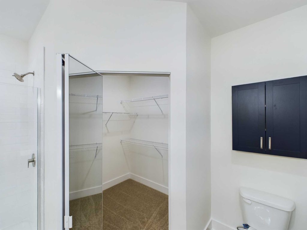 Apartments in Farragut A walk-in closet with a mirrored sliding door, white wire shelving, beige carpet, and a partial view of a bathroom with a shower, toilet, and a dark blue cabinet. Ideal for those seeking modern comfort in apartment rentals.