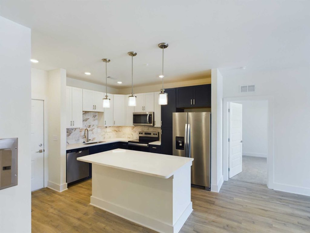 Apartments in Farragut A modern kitchen with white cabinetry, stainless steel appliances, an island with a white countertop, pendant lights, and a wood floor. A hallway with a partially open door is visible in the background, typical of upscale apartments in desirable housing markets.
