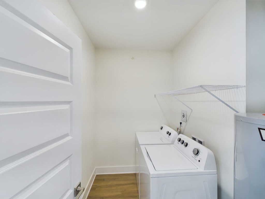 Apartments in Farragut A small laundry room in our rental properties features a white washer and dryer side by side, a wire shelf above them, a white door to the left, and light-colored walls with wooden flooring. Perfect for those seeking convenient housing solutions in modern apartments.