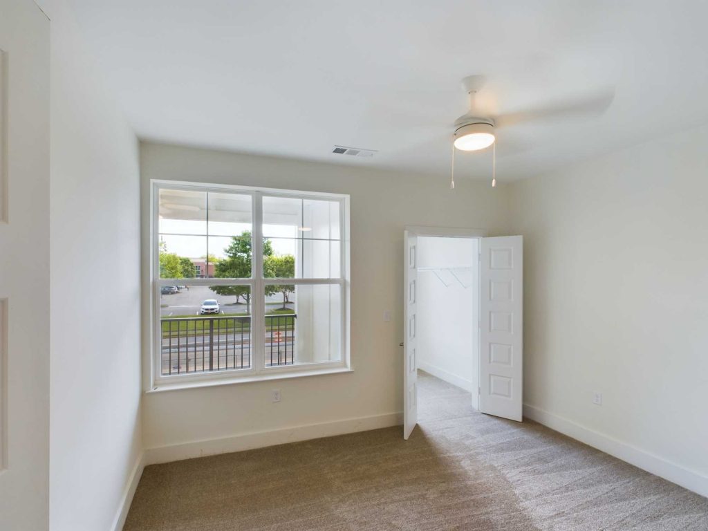 Apartments in Farragut A vacant room with beige carpet, an open closet with white double doors, a ceiling fan, and a large window overlooking a parking area and greenery—ideal for those seeking comfortable housing options in premium rental properties.