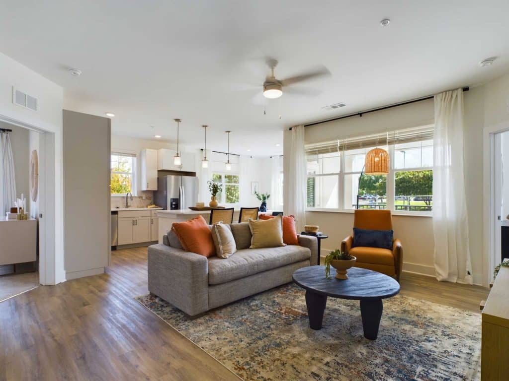 Apartments in Farragut A modern living room with a gray couch, orange pillows, a dark round coffee table, and large windows. The space features an open layout leading to a kitchen with white cabinetry and stainless steel appliances.