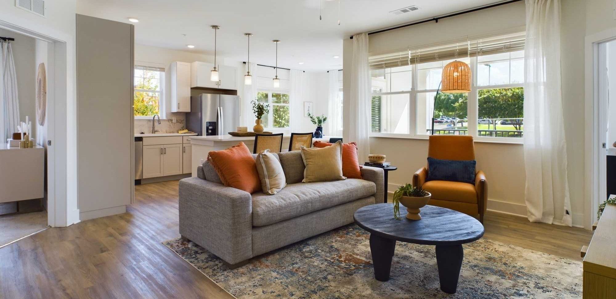 Elevate Your Apartment Living Experience In Farragut A modern living room with a gray sofa, orange cushions, and a round coffee table on a patterned rug. Large windows and a ceiling fan are visible. The kitchen is in the background. Anchor 934 Apartments in Biddle Farms Town Center