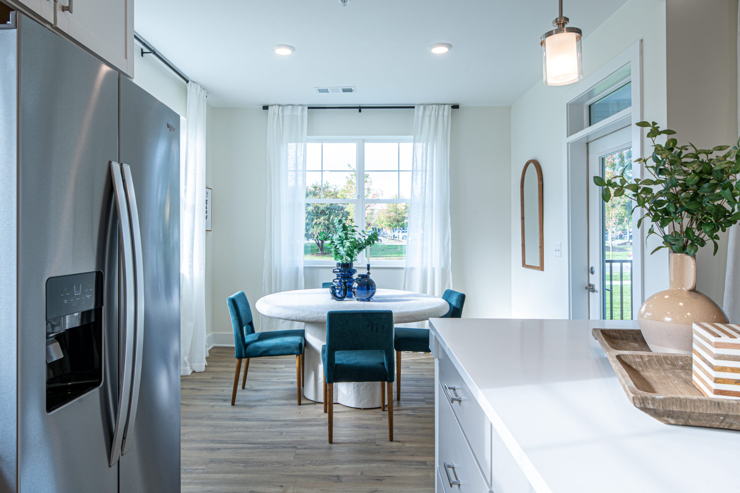 Apartments in for rent Farragut TN Modern kitchen and dining area with stainless steel refrigerator, white countertops, a round table with blue chairs, large window with sheer curtains, and decorative plants. Anchor 934 Apartments in Biddle Farms Town Center