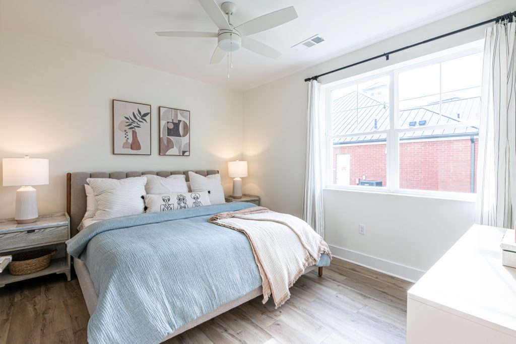 Apartments in for rent Farragut TN A bedroom with a double bed, blue and beige bedding, two bedside tables with lamps, wall art, a ceiling fan, and a window with white curtains. Anchor 934 Apartments in Biddle Farms Town Center