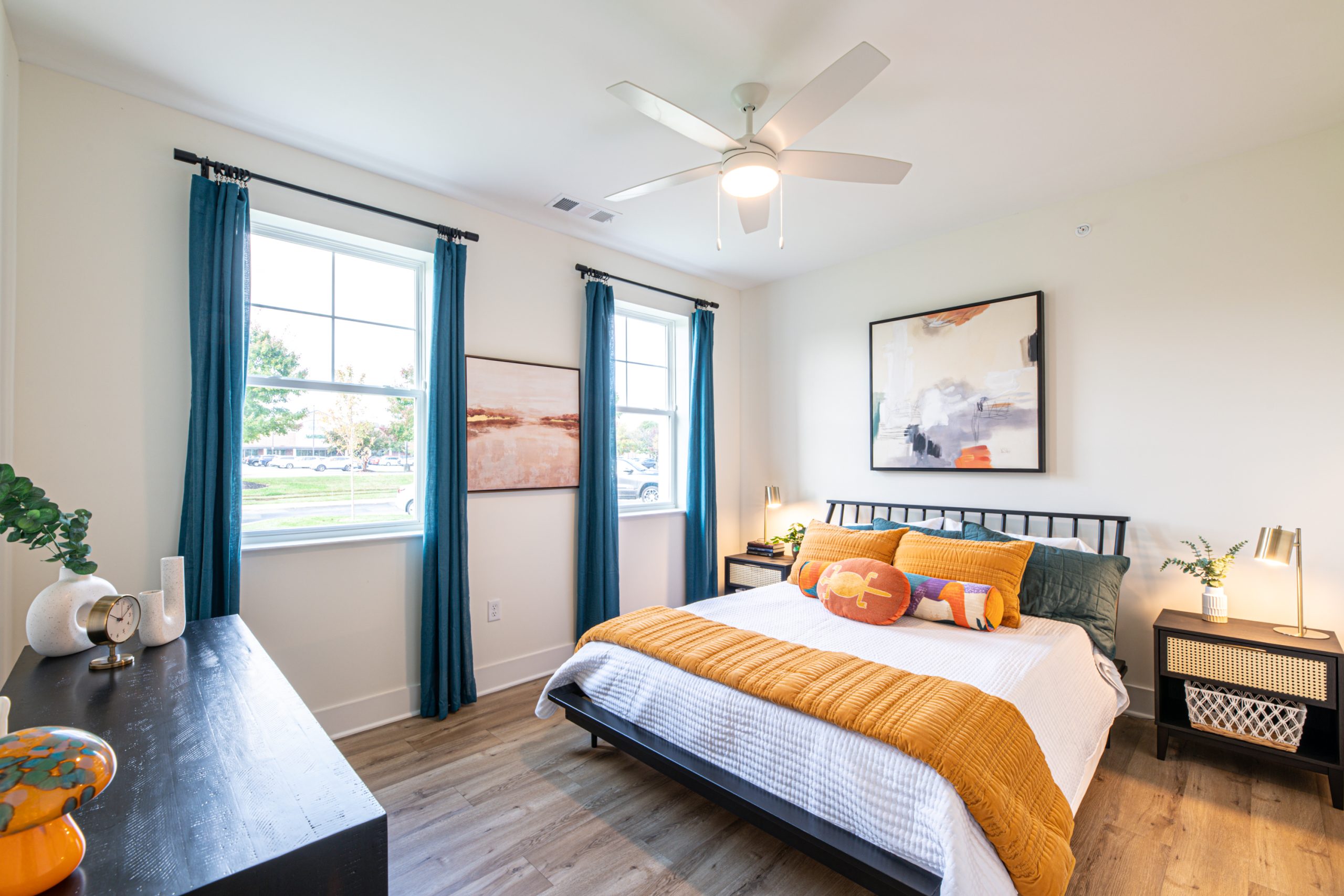 Apartments in for rent Farragut TN Modern bedroom with a bed, orange and white bedding, abstract wall art, a black dresser, two windows with blue curtains, and a ceiling fan. Anchor 934 Apartments in Biddle Farms Town Center