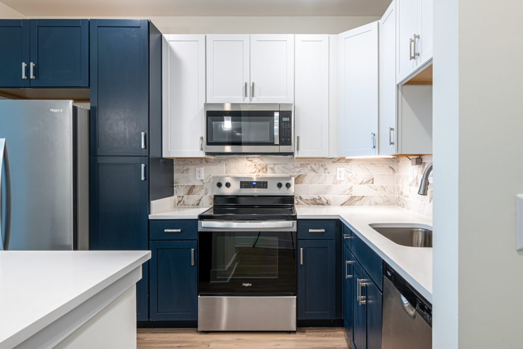 Apartments in for rent Farragut TN Modern kitchen with navy and white cabinets, stainless steel appliances including a refrigerator, oven, microwave, and dishwasher, and a marble backsplash. White countertops and wood flooring. Anchor 934 Apartments in Biddle Farms Town Center