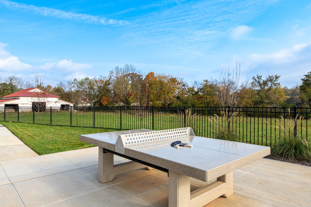 Apartments in for rent Farragut TN Concrete ping pong table with paddles on top, set in an outdoor area with trees, grass, and a black fence under a blue sky. Anchor 934 Apartments in Biddle Farms Town Center