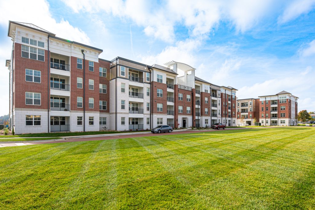 Apartments in for rent Farragut TN A wide-angle photo of a modern, multi-story apartment building with a well-kept lawn and parked cars in front, under a partly cloudy sky. Anchor 934 Apartments in Biddle Farms Town Center
