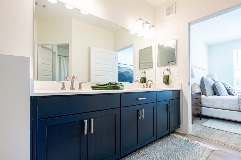 Apartments in for rent Farragut TN Modern bathroom with a large mirror, double sink vanity with dark cabinets, and a lit bedroom visible through an open door. White walls and sleek fixtures complete the setting. Anchor 934 Apartments in Biddle Farms Town Center