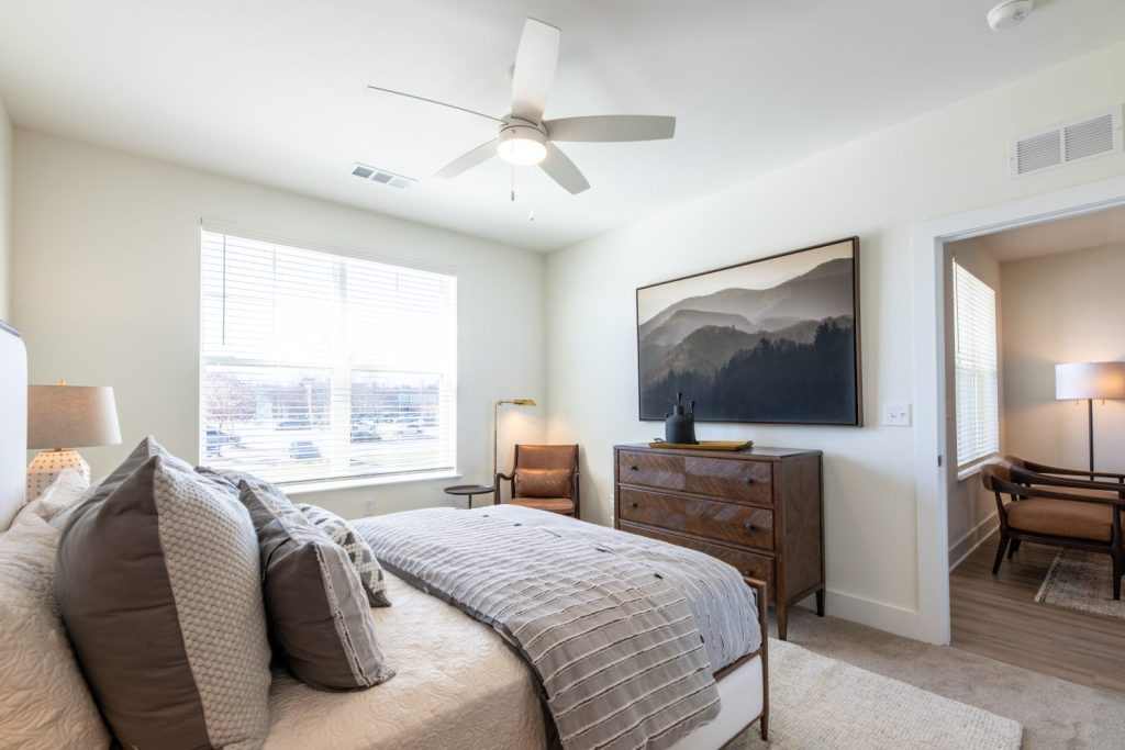 Apartments in for rent Farragut TN A bedroom with a neatly made bed, a large landscape picture, a wooden dresser, armchair, and a ceiling fan. Natural light enters through a window with blinds. Anchor 934 Apartments in Biddle Farms Town Center