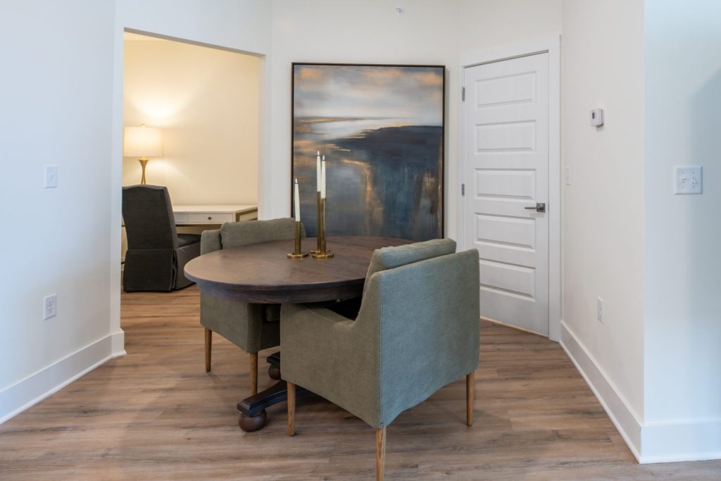 Apartments in for rent Farragut TN A small dining area with a round table, two green chairs, and a large abstract painting. A doorway opens to a room with a lamp and desk. White walls and wood flooring are visible. Anchor 934 Apartments in Biddle Farms Town Center