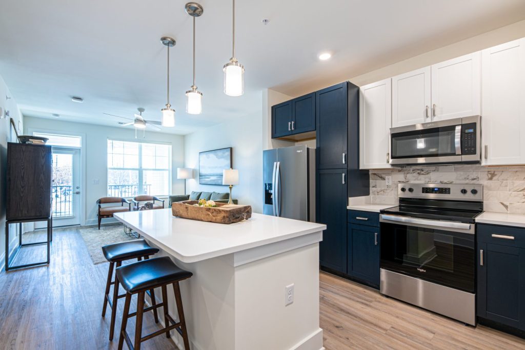 Apartments in for rent Farragut TN Modern kitchen and living area with a central island, bar stools, stainless steel appliances, pendant lights, and a seating area with a sofa near a large window. Anchor 934 Apartments in Biddle Farms Town Center