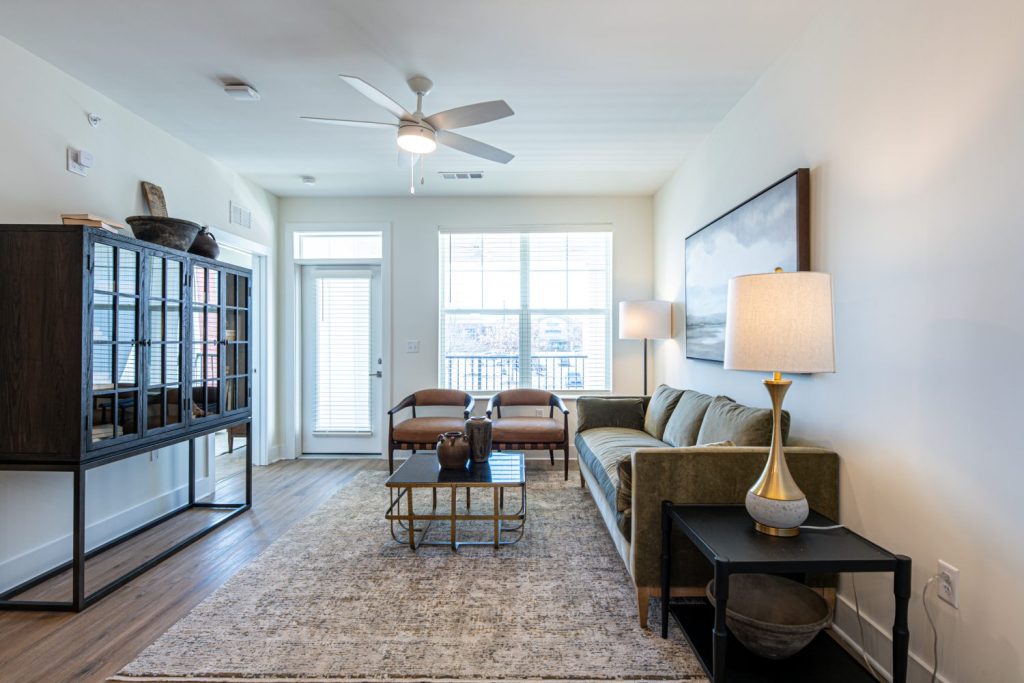 Apartments in for rent Farragut TN Modern living room with a green sofa, two brown chairs, a glass coffee table, a lamp, and a dark cabinet. Light streams in through large windows, illuminating the hardwood floor. Anchor 934 Apartments in Biddle Farms Town Center