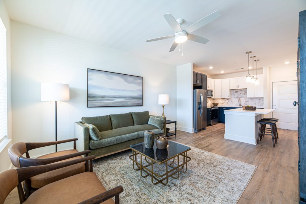 Apartments in for rent Farragut TN Modern living room with a green sofa, two wooden chairs, and a glass coffee table on a rug. A ceiling fan is above, and a kitchen with an island is visible in the background. Anchor 934 Apartments in Biddle Farms Town Center