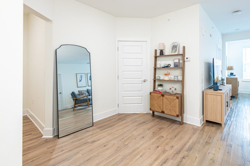 Apartments in for rent Farragut TN A modern apartment entryway with wood flooring, a large mirror, a bookshelf with decor, and a view into a living area with a chair and natural light. Anchor 934 Apartments in Biddle Farms Town Center