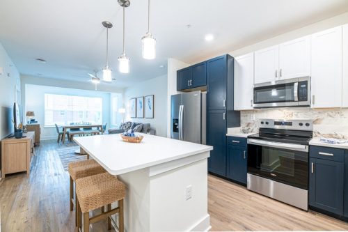 Apartments in for rent Farragut TN Modern open-concept kitchen with blue and white cabinets, stainless steel appliances, an island with two wicker stools, and a view of the dining and living area in the background. Anchor 934 Apartments in Biddle Farms Town Center