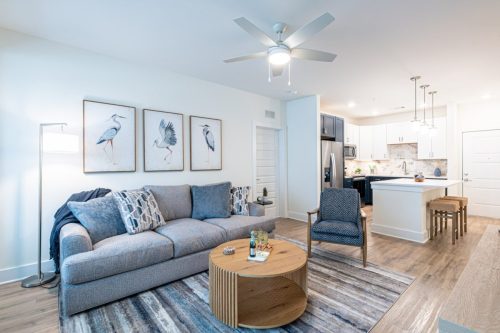 Apartments in for rent Farragut TN Modern living room with a gray sofa, two patterned chairs, a round wooden coffee table, and wall art of birds, adjacent to an open kitchen with barstools. Anchor 934 Apartments in Biddle Farms Town Center