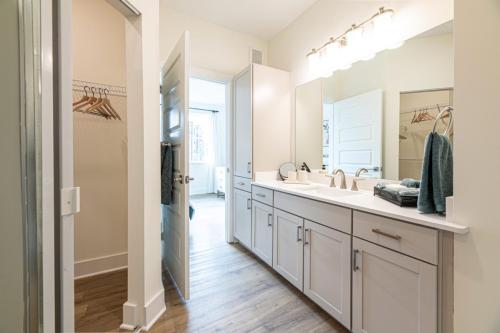 Apartments in for rent Farragut TN Modern bathroom with double sinks, large mirror, and white cabinets. The room has wooden flooring, and an open door leads to a closet with hangers and a glimpse of a bedroom. Anchor 934 Apartments in Biddle Farms Town Center