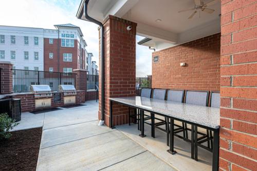 Apartments in for rent Farragut TN Outdoor patio with a long granite table and barstools, adjacent to two stainless steel grills. Red brick walls and fence, with buildings in the background. Anchor 934 Apartments in Biddle Farms Town Center