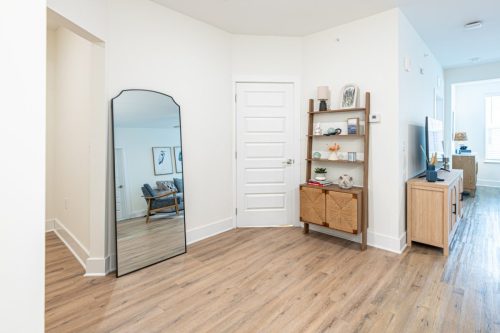Apartments in for rent Farragut TN A modern apartment entryway with wood flooring, a large mirror, a bookshelf with decor, and a view into a living area with a chair and natural light. Anchor 934 Apartments in Biddle Farms Town Center