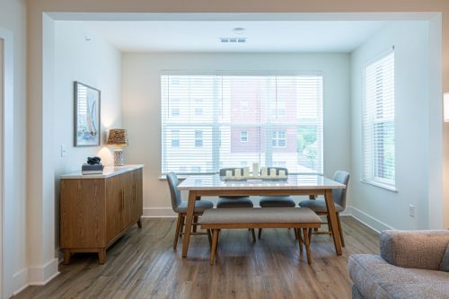 Apartments in for rent Farragut TN A modern dining area with a wooden table, six chairs, a sideboard, lamp, framed artwork, and large windows letting in natural light. Anchor 934 Apartments in Biddle Farms Town Center