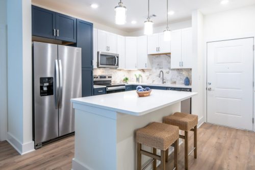 Apartments in for rent Farragut TN Modern kitchen with stainless steel appliances, white and navy cabinets, marble backsplash, an island with two wicker stools, and wooden floors. Anchor 934 Apartments in Biddle Farms Town Center