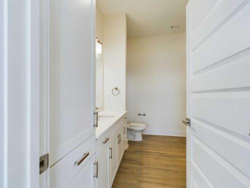 Apartments in for rent Farragut TN A hallway bathroom with a white vanity, countertop, and mirror on the left, a wall-mounted light above, and a toilet in the background. The floor is wood and the walls are light-colored. Anchor 934 Apartments in Biddle Farms Town Center