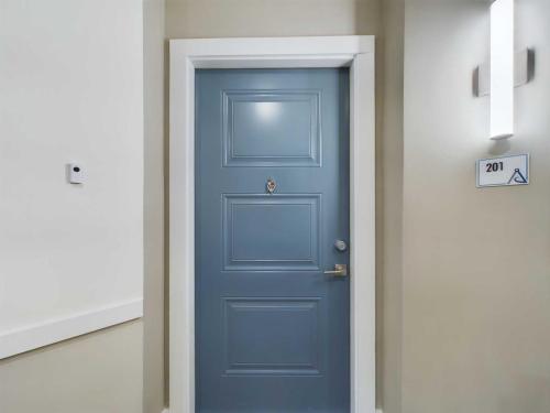 Three Bedroom Apartment Sailor Apt. 1-201 Apartments in for rent Farragut TN A blue door labeled 201 with a handle and peephole is centered between beige walls and white trim. A modern wall lamp is on the right. Anchor 934 Apartments in Biddle Farms Town Center