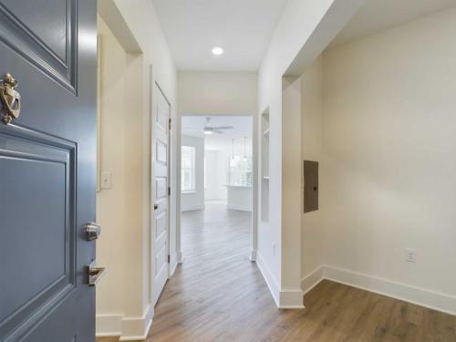 Apartments in for rent Farragut TN View of a modern, empty hallway with light wood flooring, leading to an open living area with large windows. A blue front door is partially open in the foreground. Anchor 934 Apartments in Biddle Farms Town Center